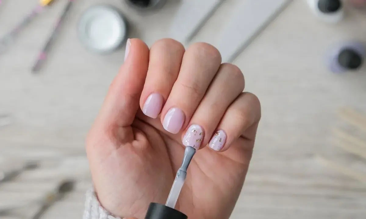 Dłoń z paznokciami w pastelowym różu, jedna ręka nakłada lakier. W tle akcesoria do **nailprep**.