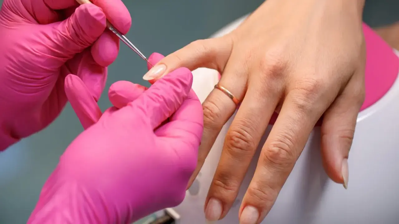 Dłonie w różowych rękawiczkach wykonują precyzyjny manicure, przygotowując paznokcie na letnie kolory.
