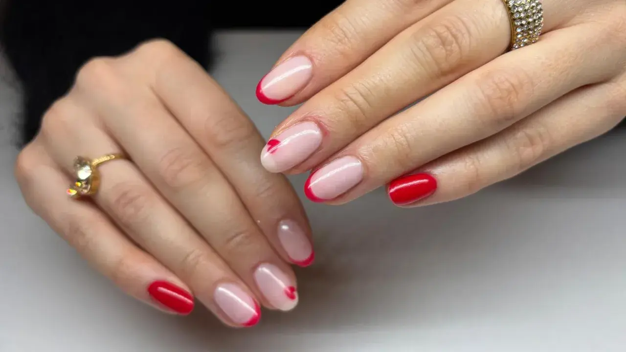 Dłonie z paznokciami w stylu czerwony french. Na niektórych paznokciach subtelne serduszka. Złote pierścionki dodają blasku.