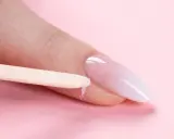Delikatne baby boomer nails w trakcie stylizacji. Drewniany patyczek usuwa nadmiar produktu z okolicy paznokcia.