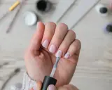 Dłoń z paznokciami w pastelowym różu, jedna ręka nakłada lakier. W tle akcesoria do **nailprep**.
