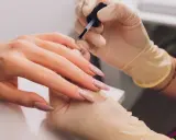 Dłonie w rękawiczkach nakładają lakier na paznokcie, tworząc delikatny french.