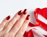 Dłoń z paznokciami neonowymi czerwonymi, błyszczącymi jak drogocenne kamienie, obok lśniącej, czerwonej wstążki.