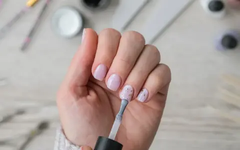 Dłoń z paznokciami w pastelowym różu, jedna ręka nakłada lakier. W tle akcesoria do **nailprep**.