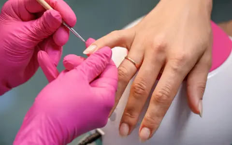 Dłonie w różowych rękawiczkach wykonują precyzyjny manicure, przygotowując paznokcie na letnie kolory.