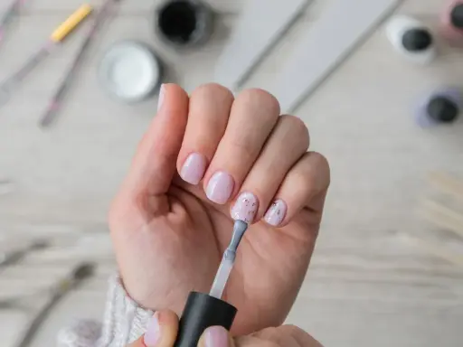 Dłoń z paznokciami w pastelowym różu, jedna ręka nakłada lakier. W tle akcesoria do **nailprep**.