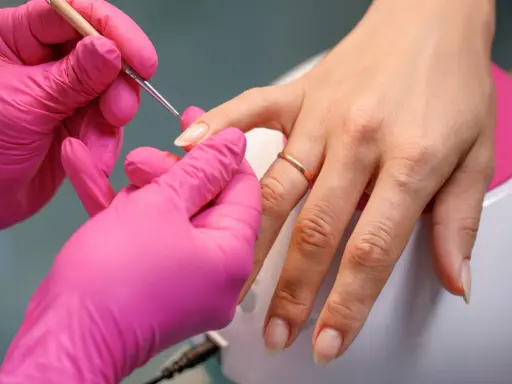Dłonie w różowych rękawiczkach wykonują precyzyjny manicure, przygotowując paznokcie na letnie kolory.