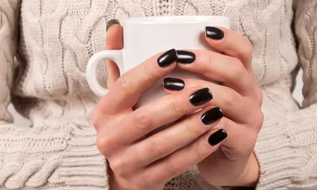 Dłonie w beżowym swetrze trzymają biały kubek. Lśniące, czarne paznokcie dodają stylizacji elegancji.