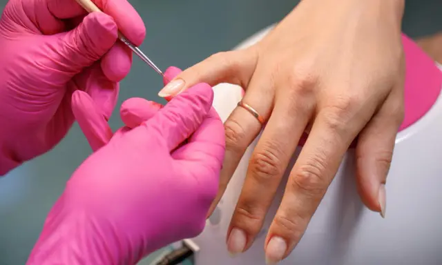 Dłonie w różowych rękawiczkach wykonują precyzyjny manicure, przygotowując paznokcie na letnie kolory.