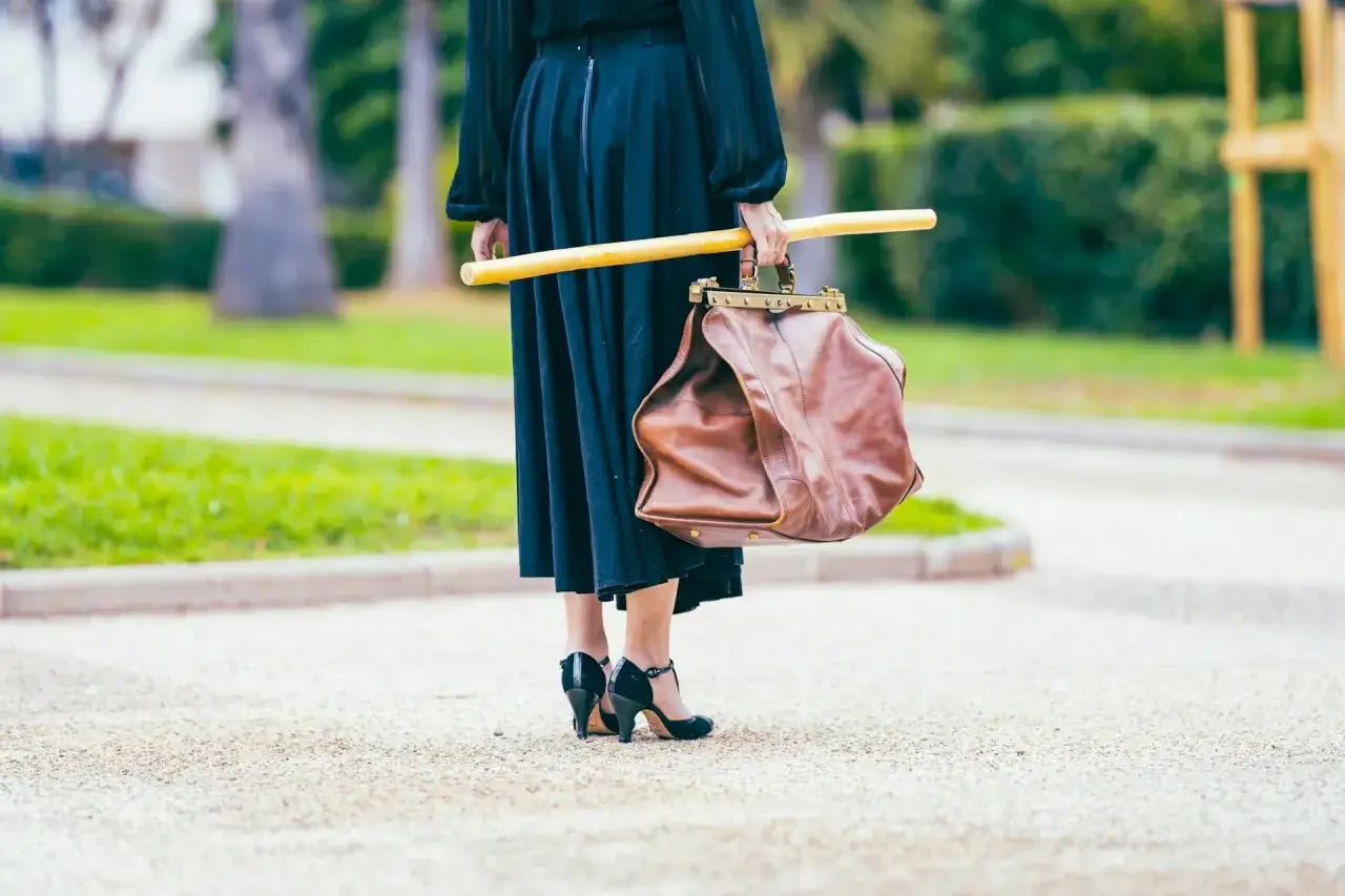 Elegancka kobieta w czarnej sukni i czarnych szpilkach trzyma brązową torbę i żółty kij. Jej wiosenne paznokcie french idealnie pasują do stylizacji.